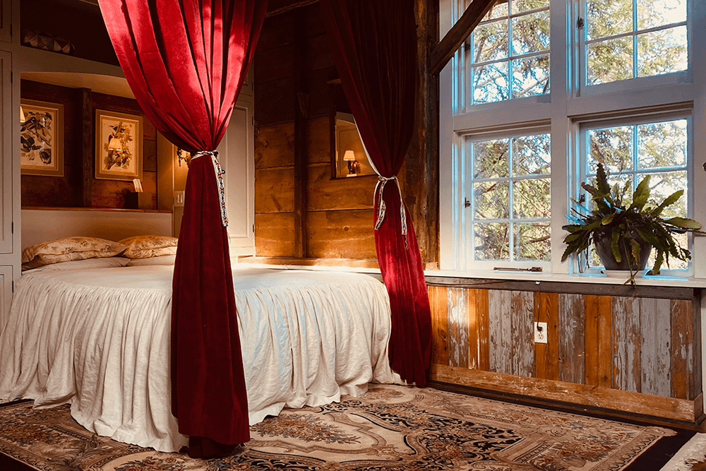 A cozy bedroom with a bed draped in white fabric, framed by red curtains, and a large window that lets in natural light.