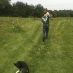 man walking in grassy meadow with black dog behind him and small whuite dog over his shoulder