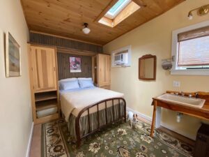 Queen bed with cream bedspread and pillows with shams, wood cabinets on both sides with shelves, sink in wood stand, dog on carpet